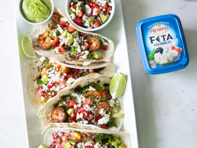 Summer shrimp tacos with strawberry avocado feta salsa made with Président Feta Crumbles, served with green crema and fresh lime on a white platter.