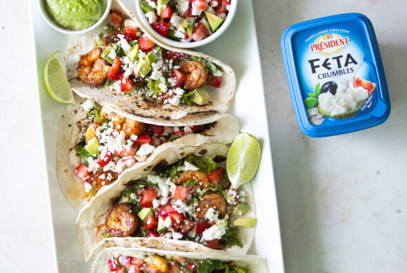 Summer shrimp tacos with strawberry avocado feta salsa made with Président Feta Crumbles, served with green crema and fresh lime on a white platter.