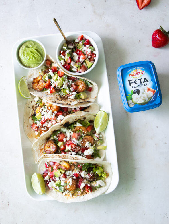 Summer shrimp tacos with strawberry avocado feta salsa made with Président Feta Crumbles, served with green crema and fresh lime on a white platter.